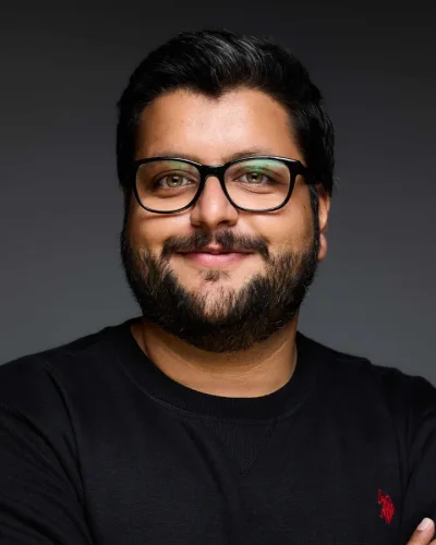 Abdul Hannan Shaikh, a bearded man in dark-framed glasses and a black jumper, arms folded, against a grey studio backdrop