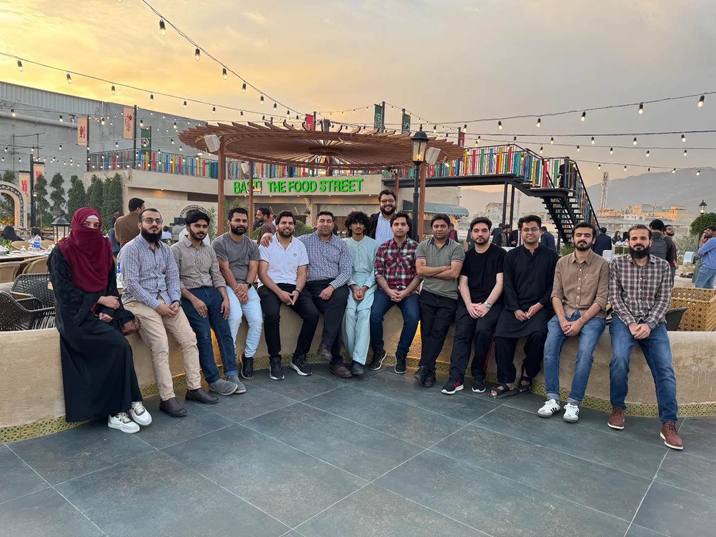The APIMatic team at an iftar dinner, fourteen people seated along a low wall on a restaurant rooftop at sunset with string lights overhead